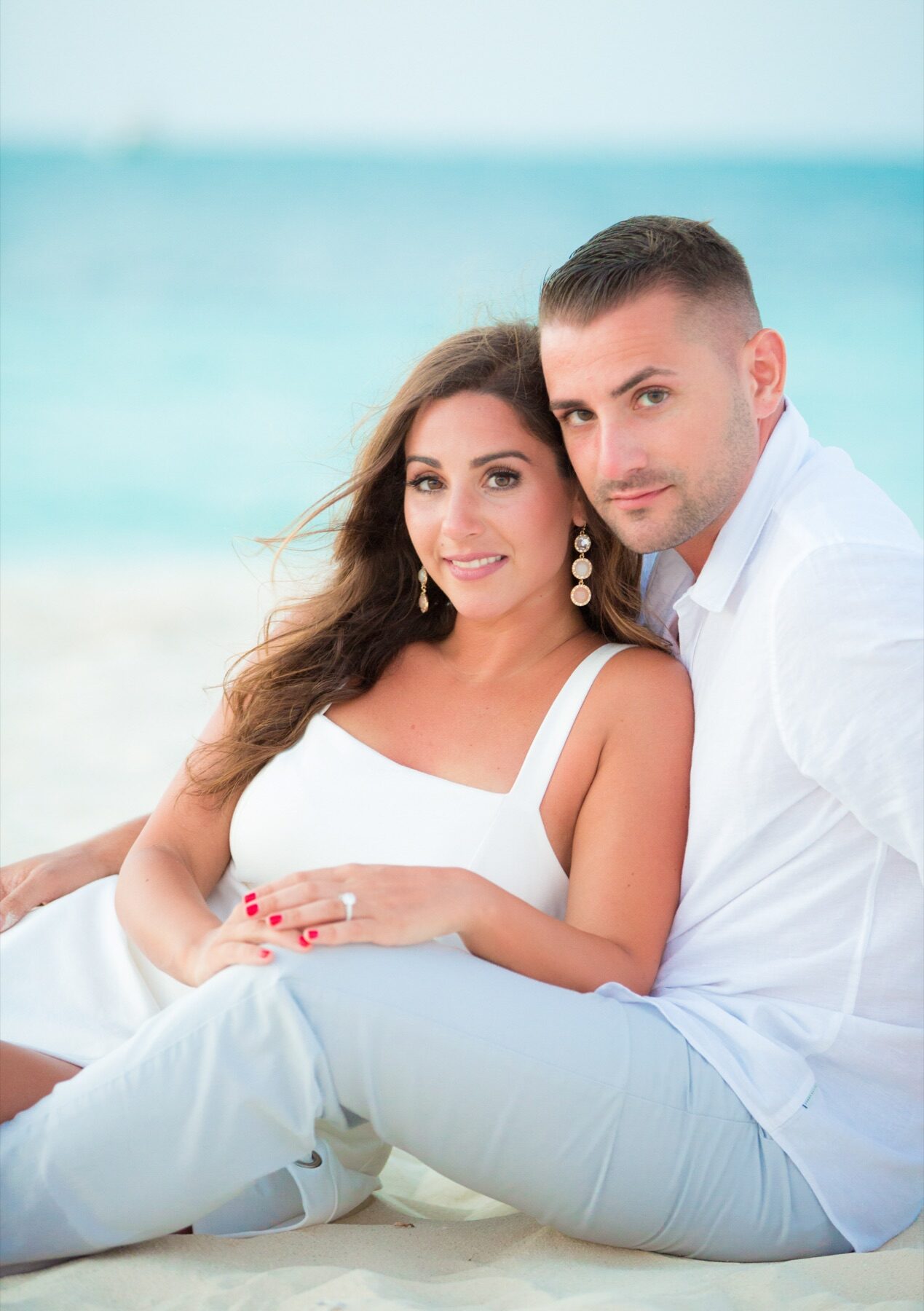 Couple sitting close on the beach smiling at the camera, turquoise water behind them