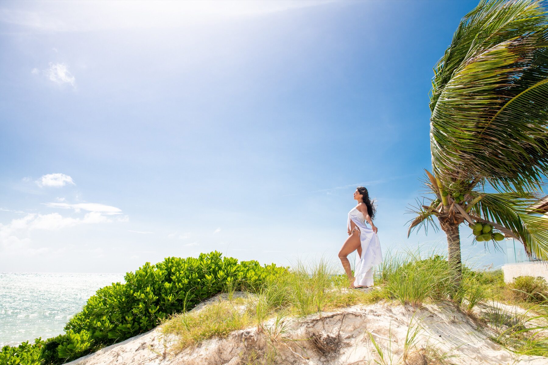 Woman in a flowing white dress standing on a dune beside a leaning palm tree