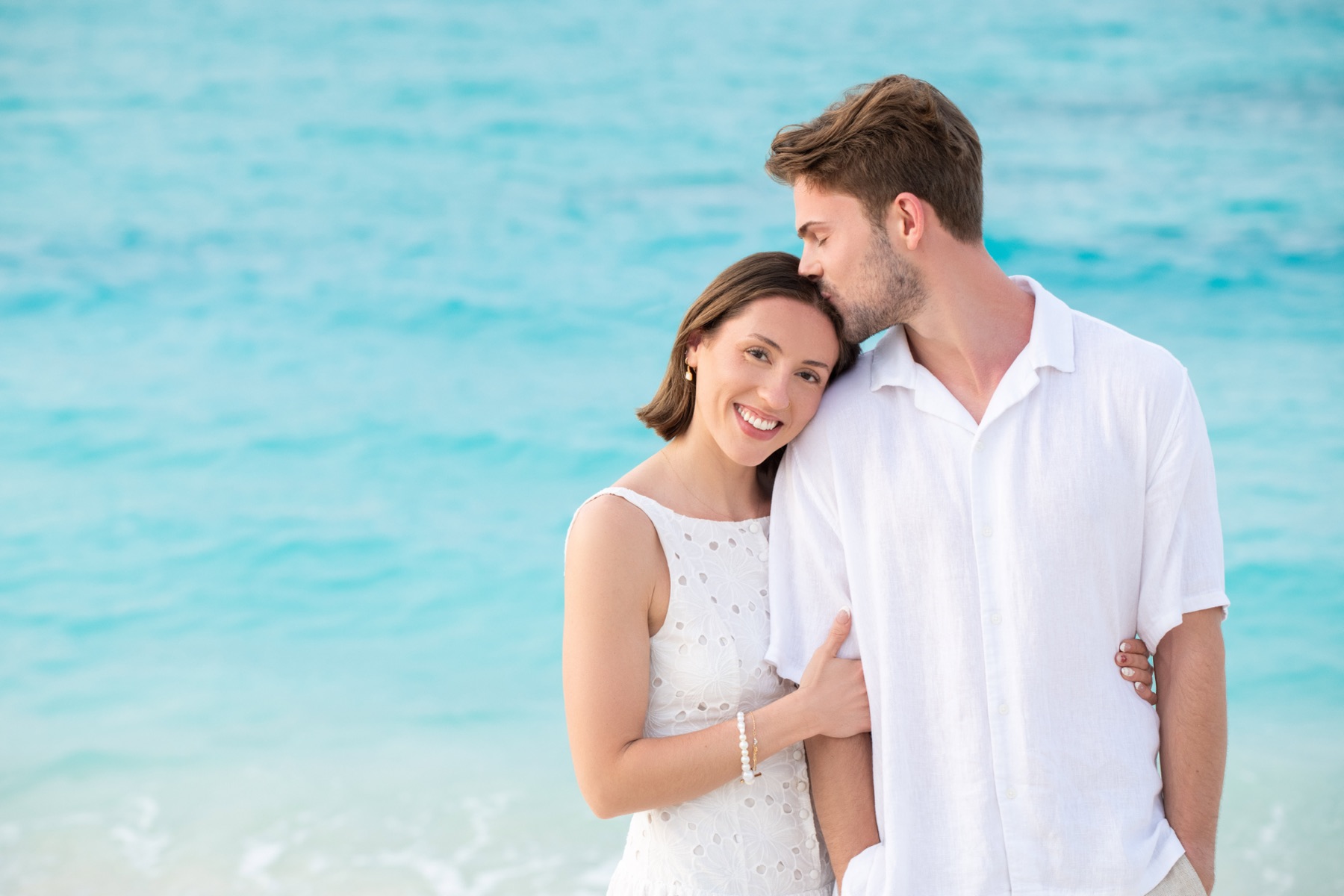 Man kissing his partner's forehead by turquoise water in matching white outfits