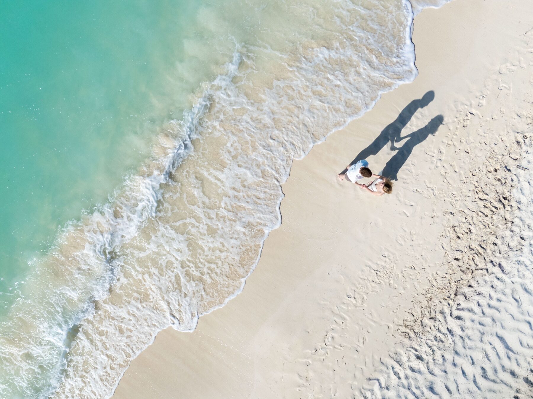 Aerial view of a couple holding hands on the shoreline, long shadows stretching across the sand