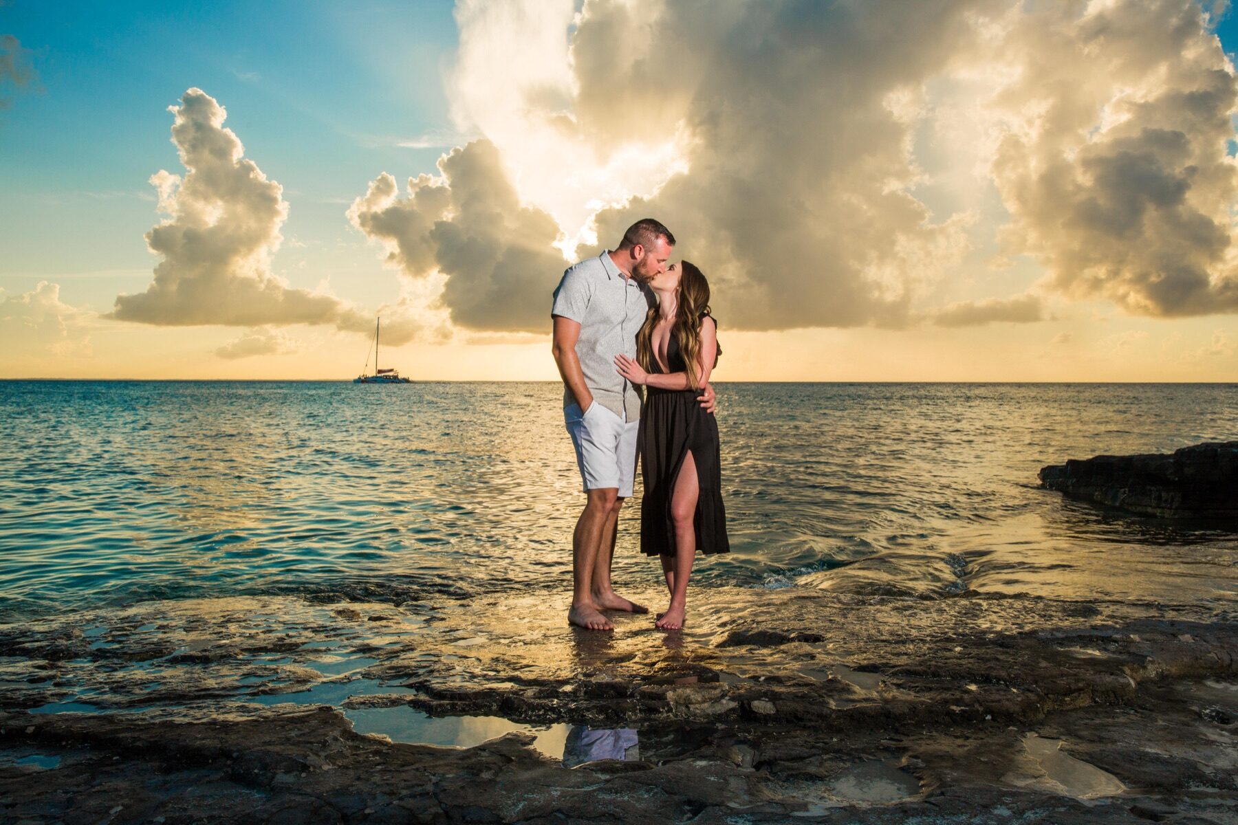 Couple kissing on rocks at sunset with a sailboat on the horizon