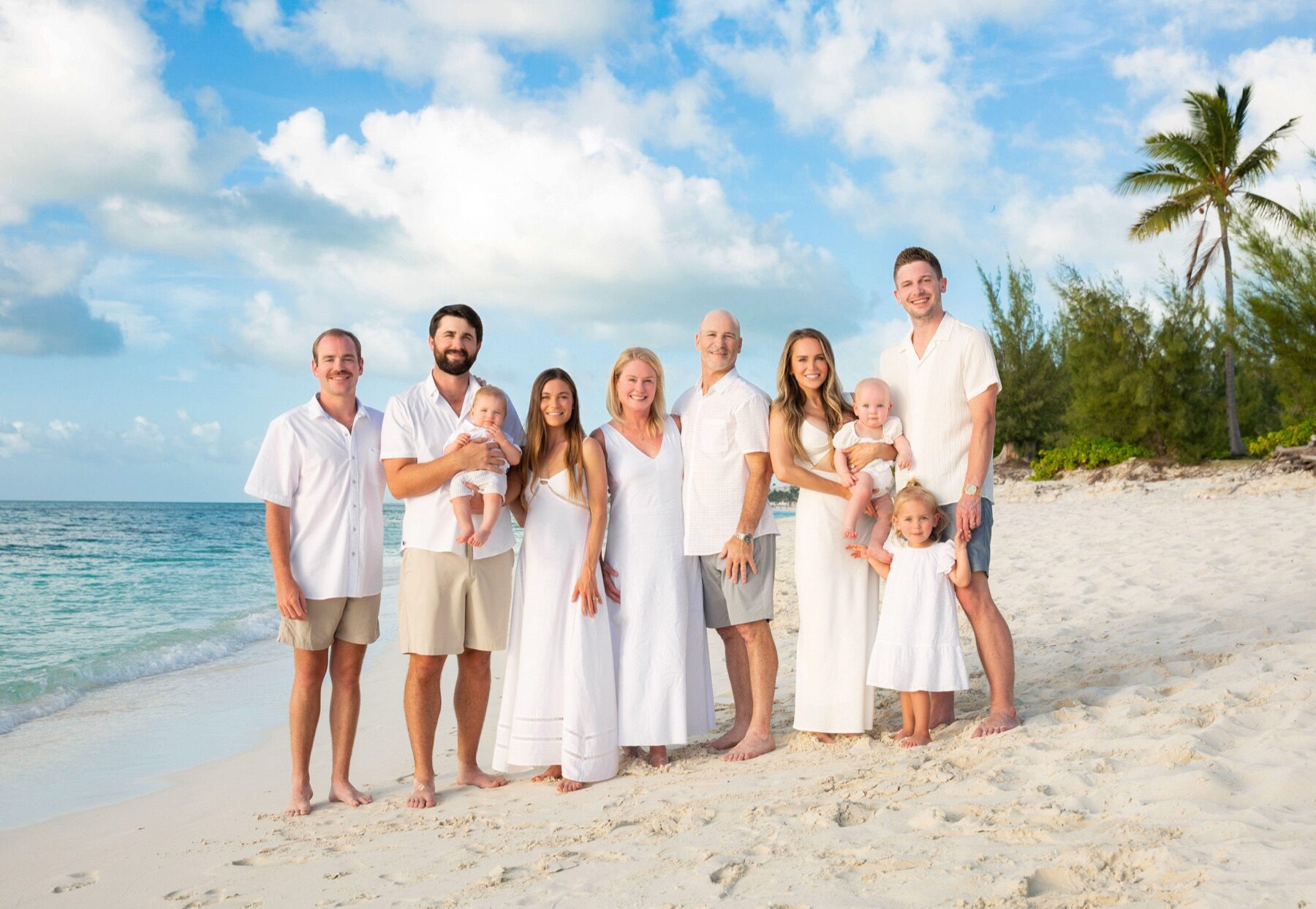 Extended family of nine in white standing together on the sand with a palm tree