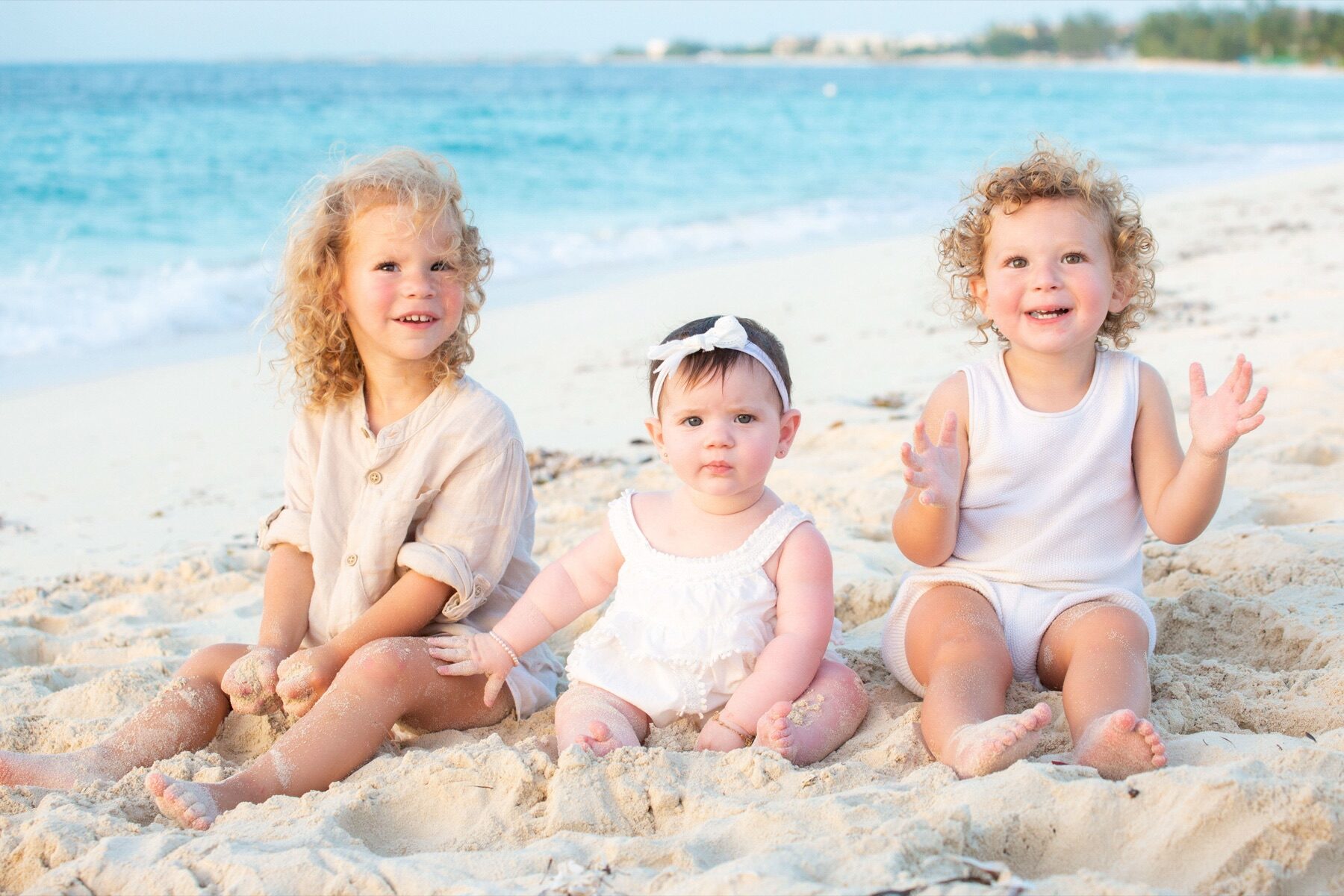 Three young children sitting together in the sand, one holding up her hands