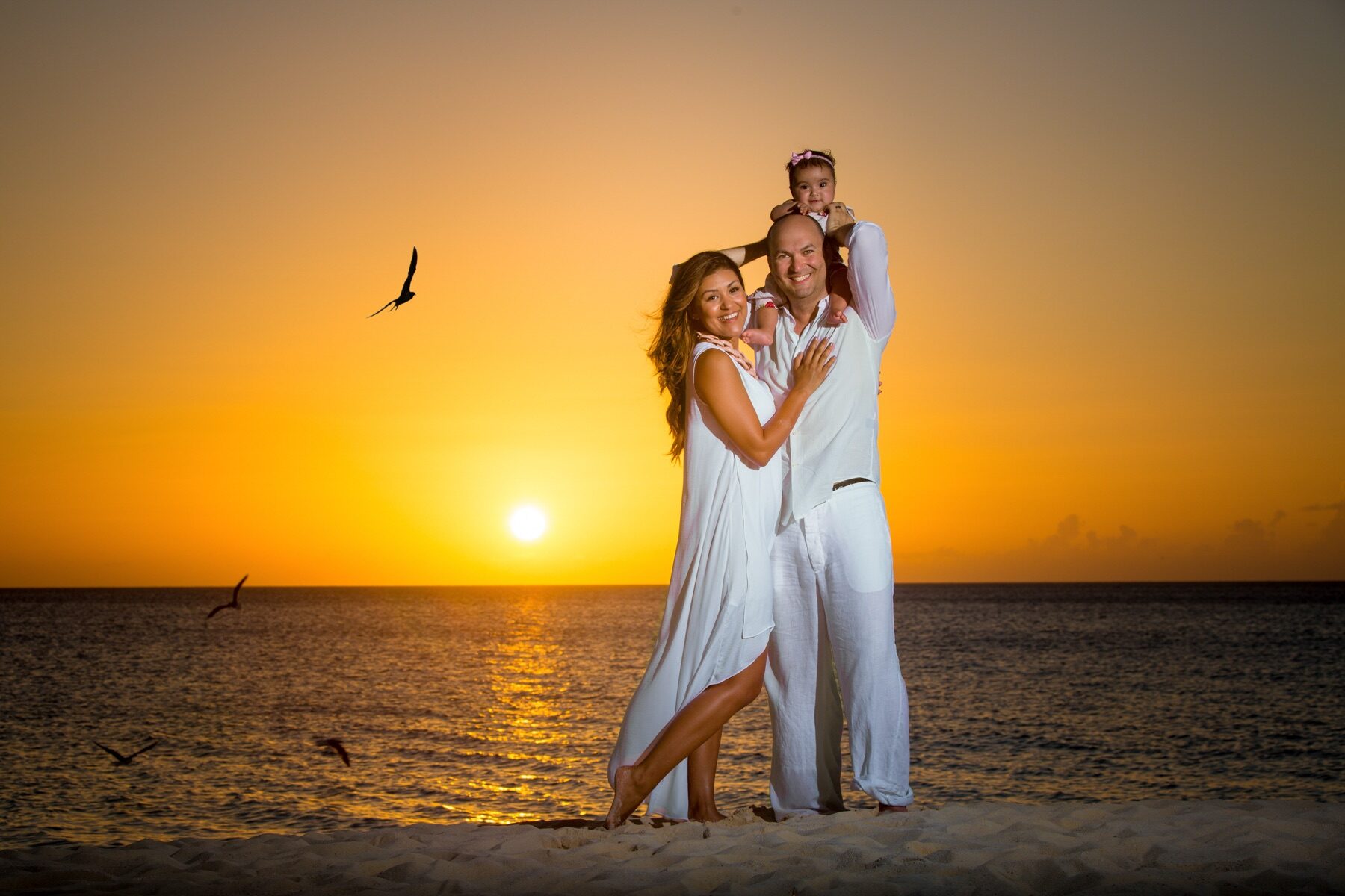 Family embracing on the beach as a toddler sits on her father's shoulders at sunset