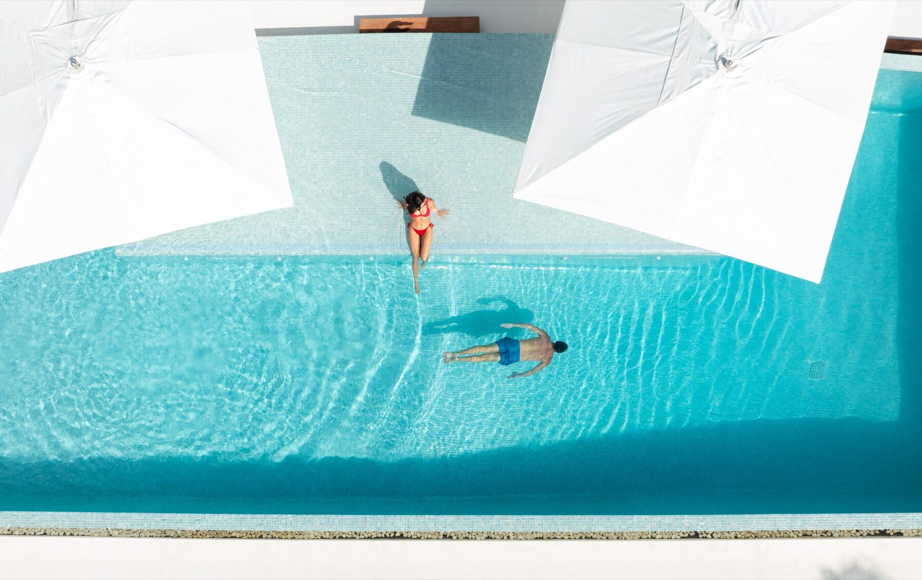 Aerial view of two swimmers floating in a pool shaded by white umbrellas