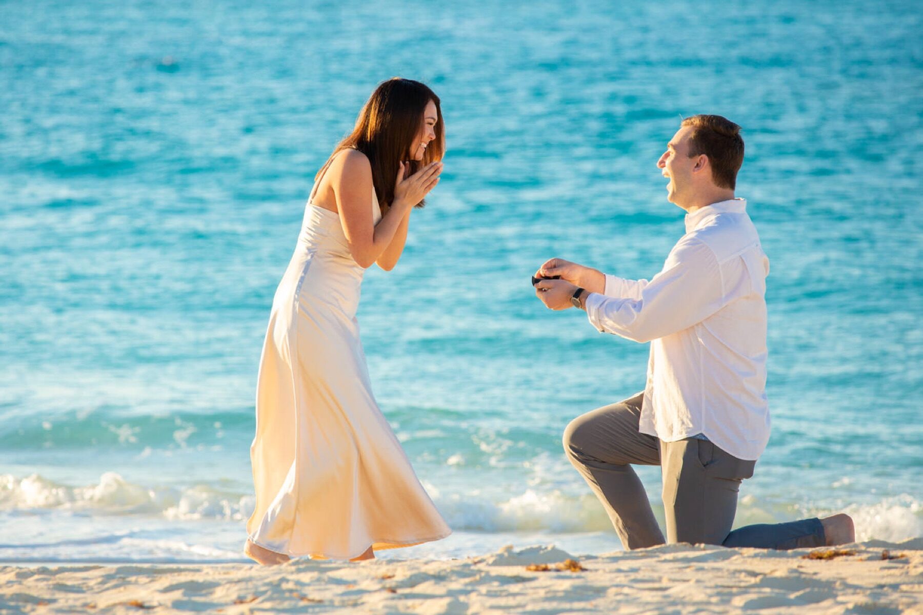 Man kneeling to propose on the beach as his partner covers her mouth in surprise