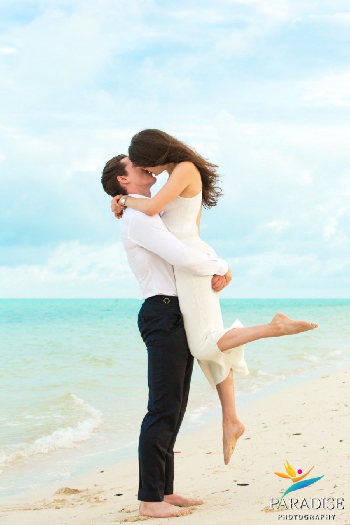 Man lifting and kissing his fiancee, her leg kicked up, on a bright beach