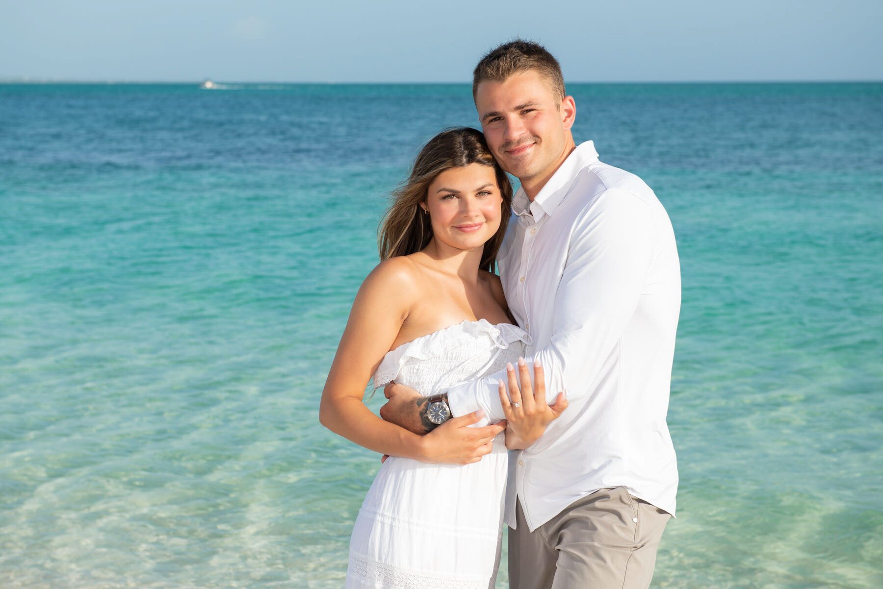 Newly engaged couple in white embracing by calm turquoise water