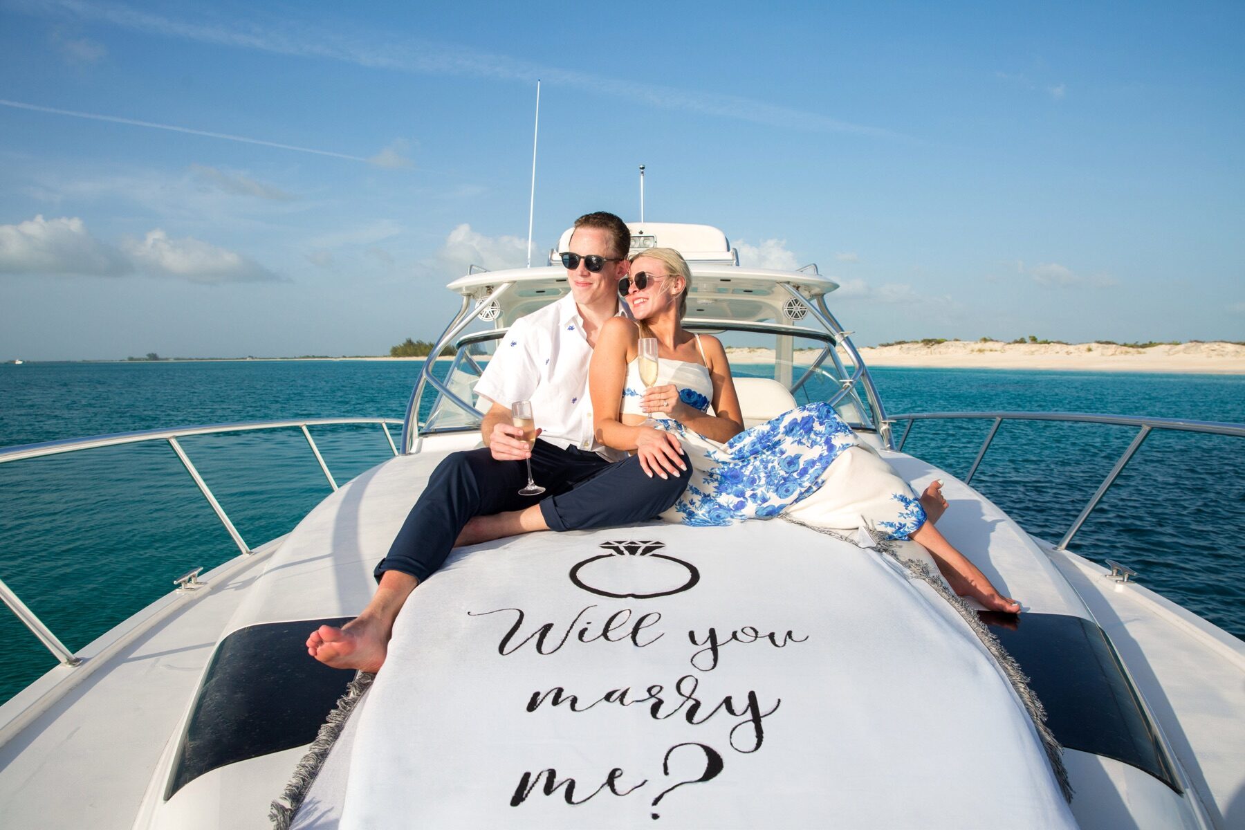Couple toasting with champagne on a yacht bow beside a will you marry me sign