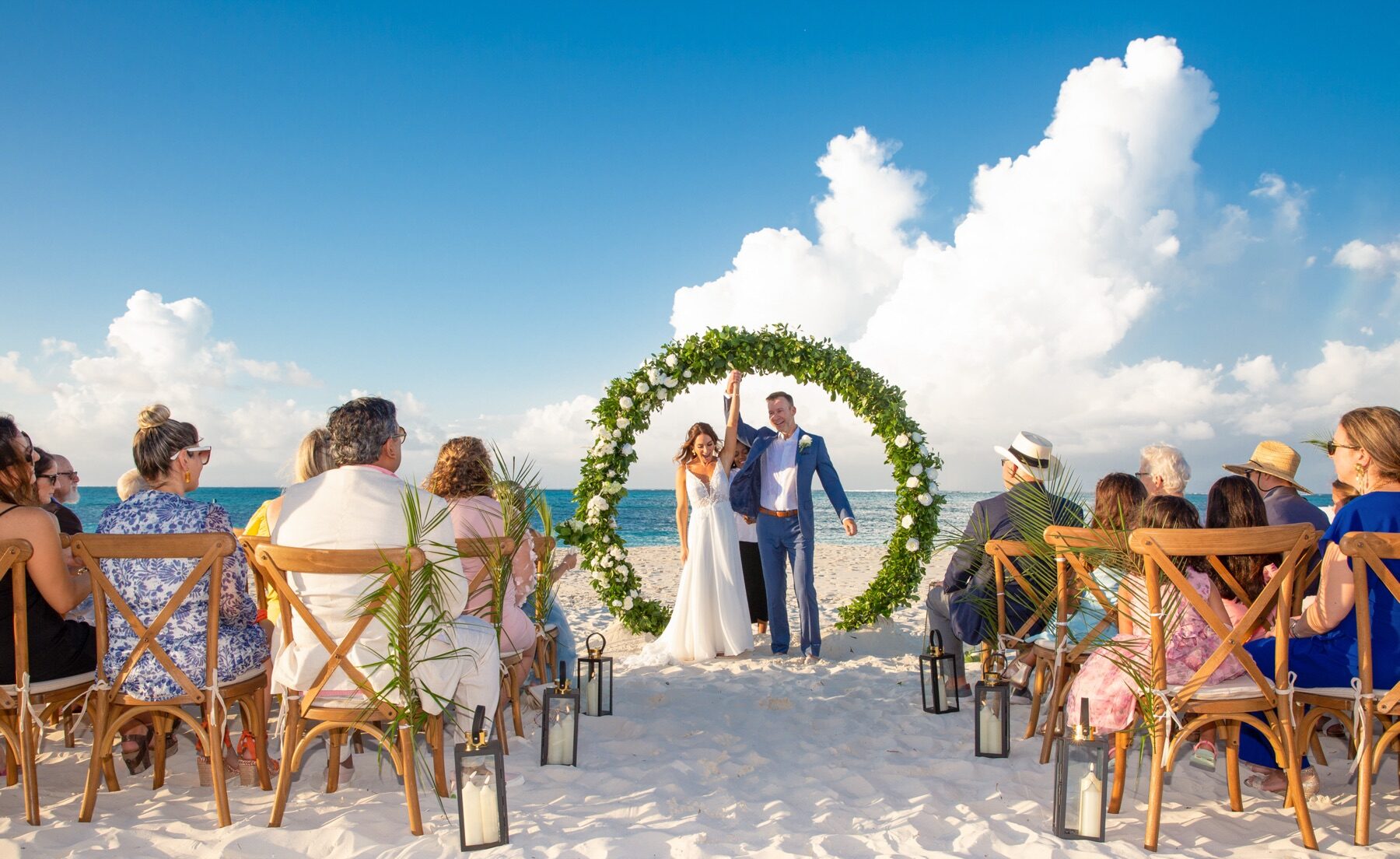 Newlyweds raising joined hands under a round greenery arch as guests watch