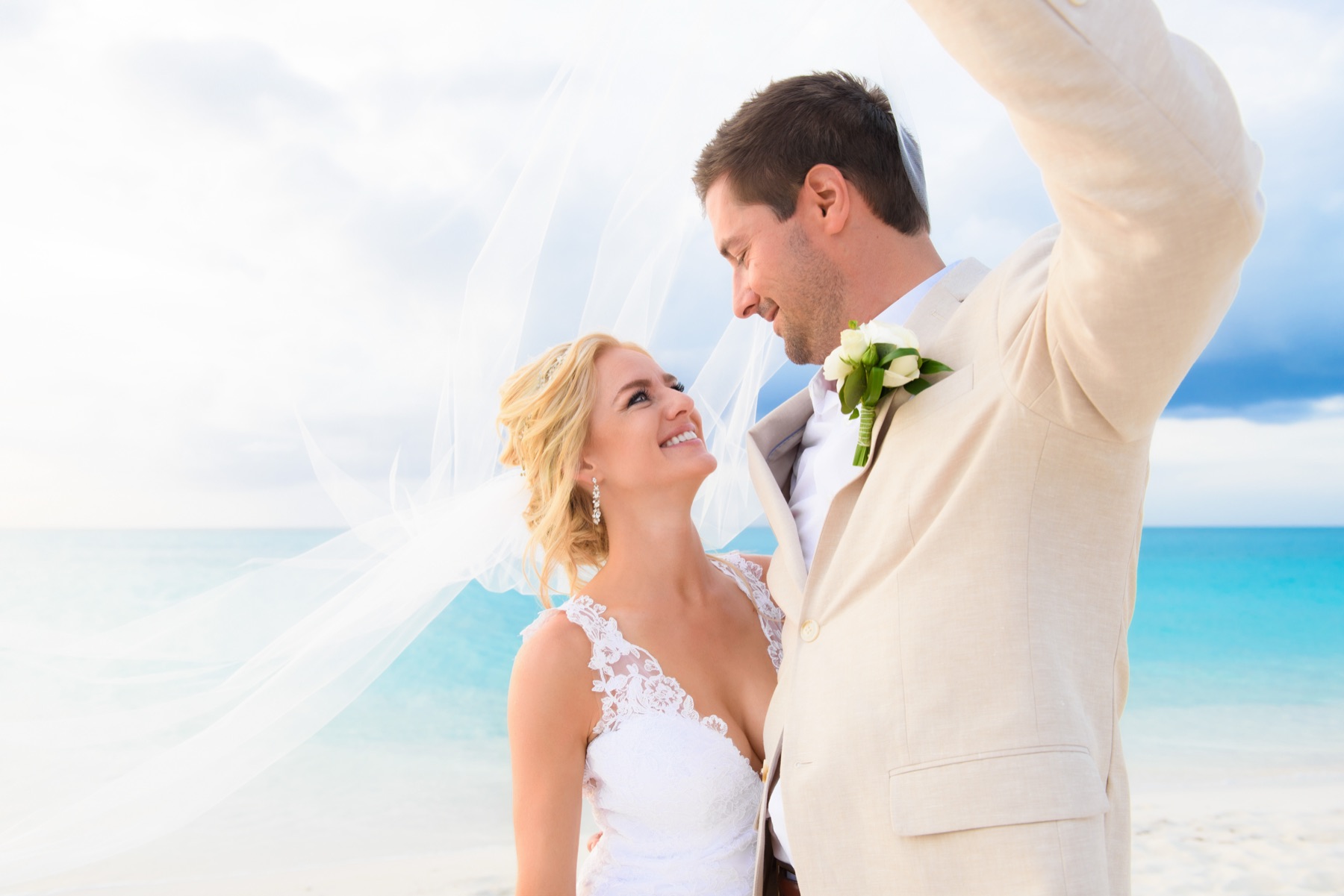 Bride laughing as her veil blows across the groom's face in the ocean breeze