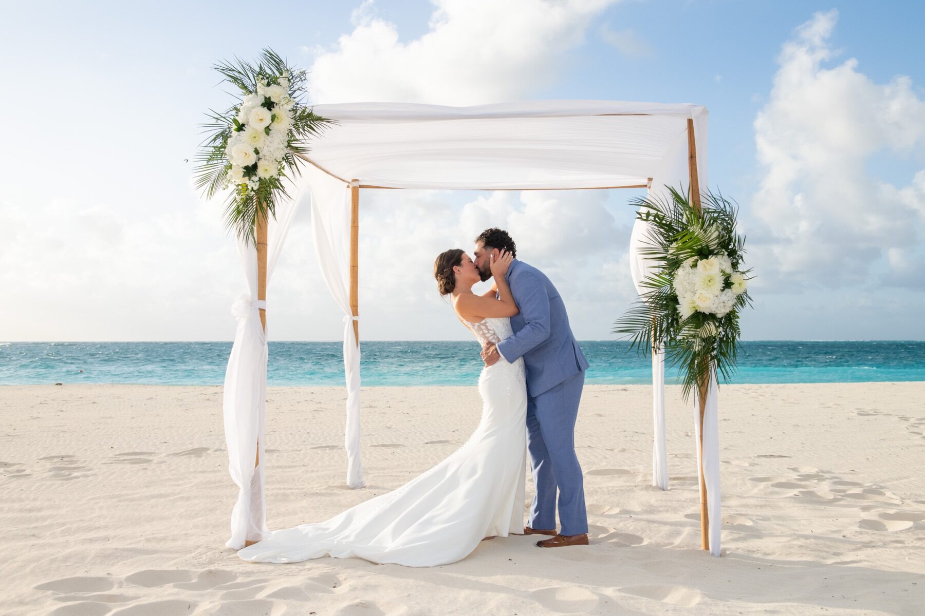 Couple kissing under a draped canopy decorated with white florals and palm fronds