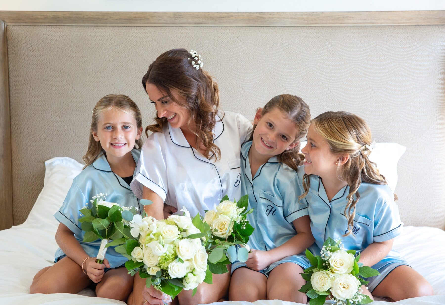 Bride in satin pajamas smiling with three flower girls holding bouquets on a bed