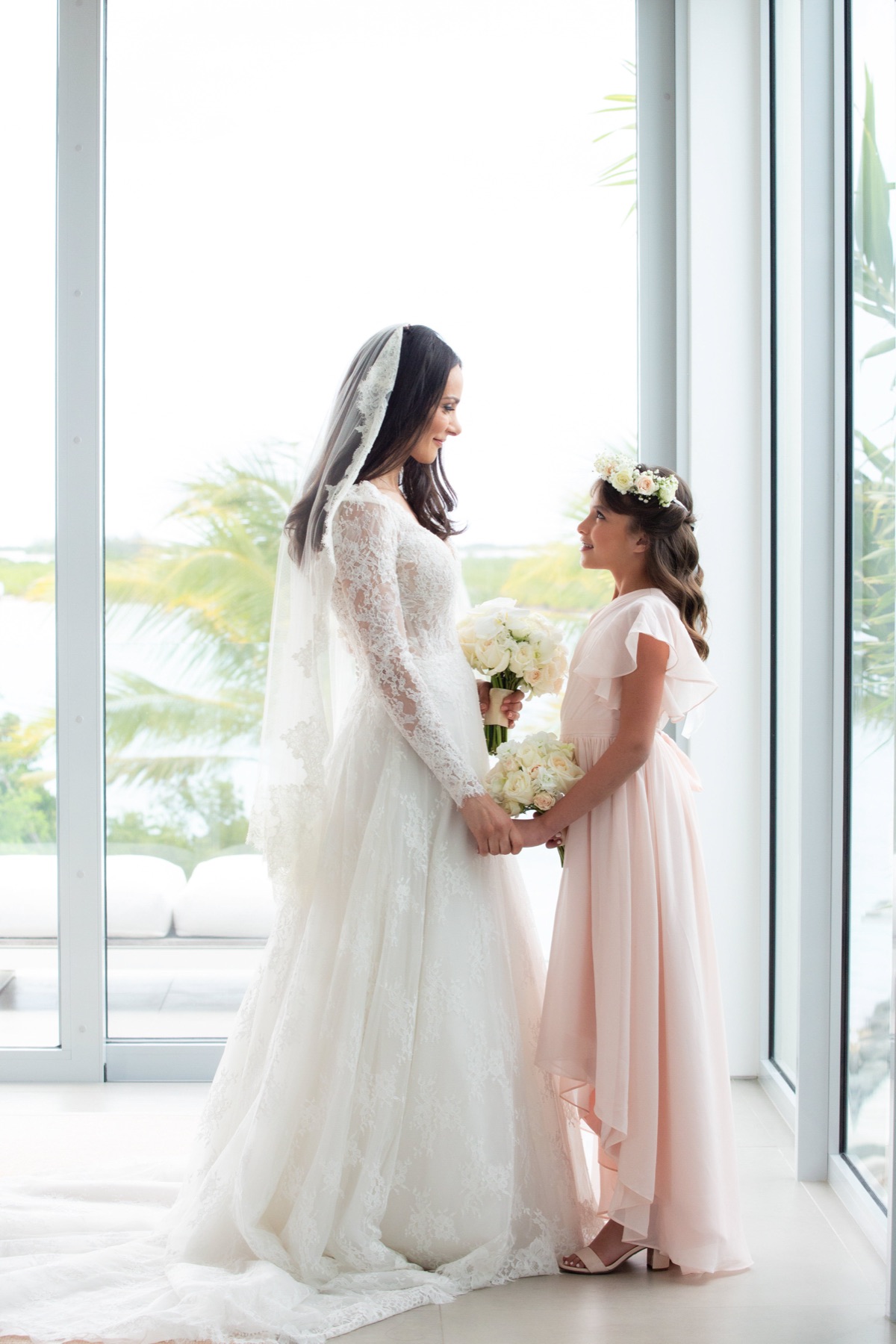Bride in lace veil holding hands with a flower girl in a pink dress by tall windows