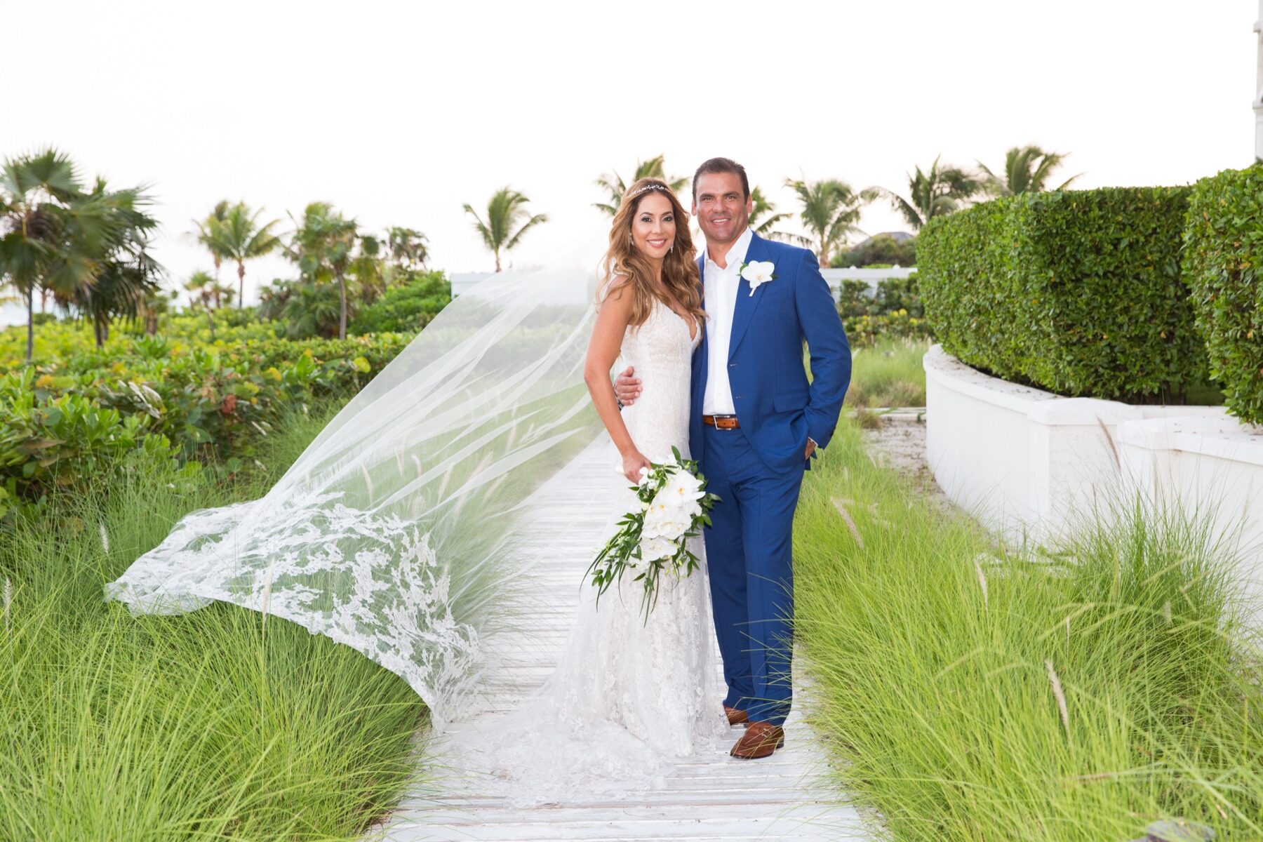 Bride and groom in blue suit strolling a Turks and Caicos boardwalk as her veil flies