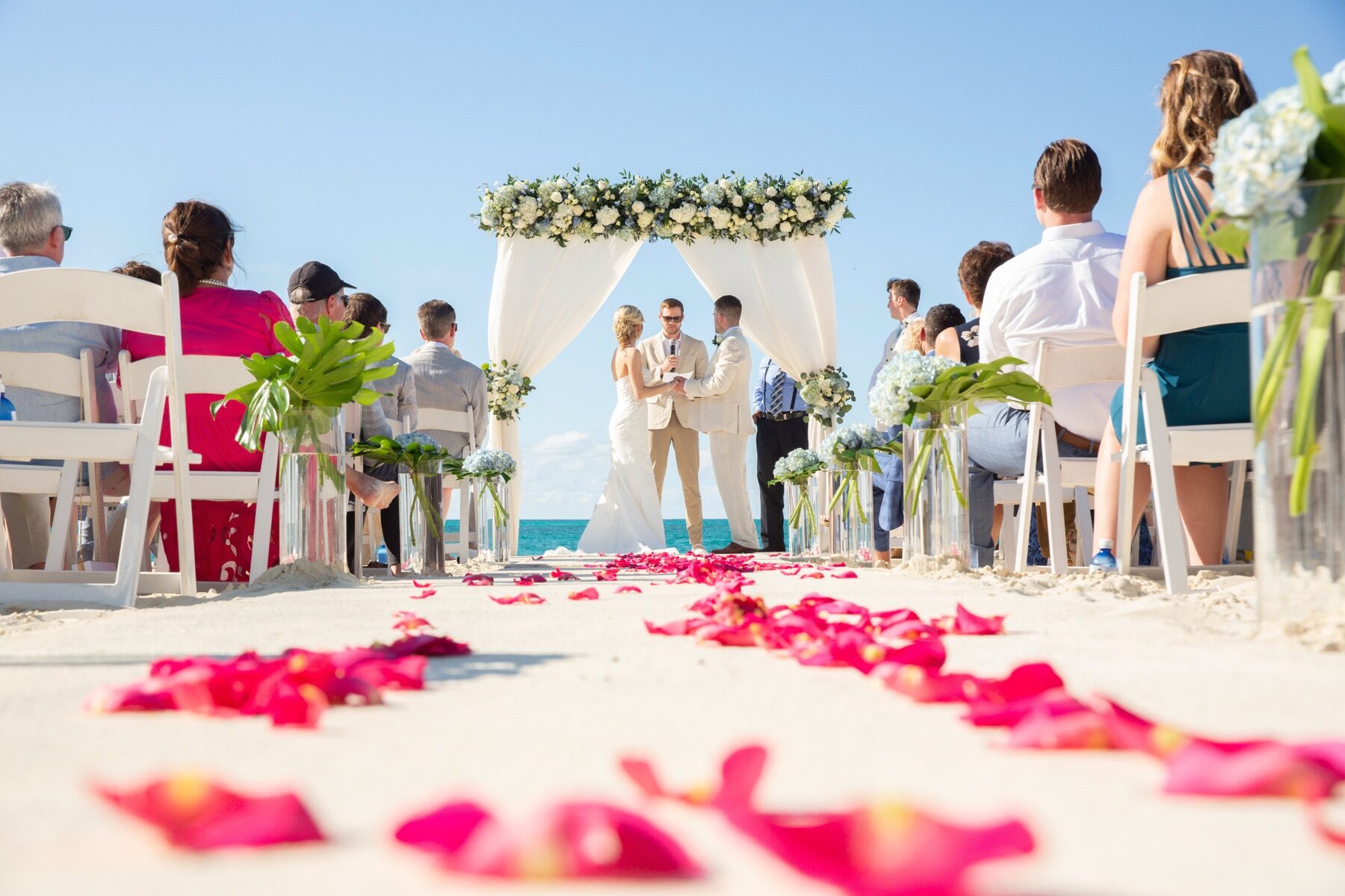 Officiant marrying a couple under a floral arch with pink petals scattered on the aisle