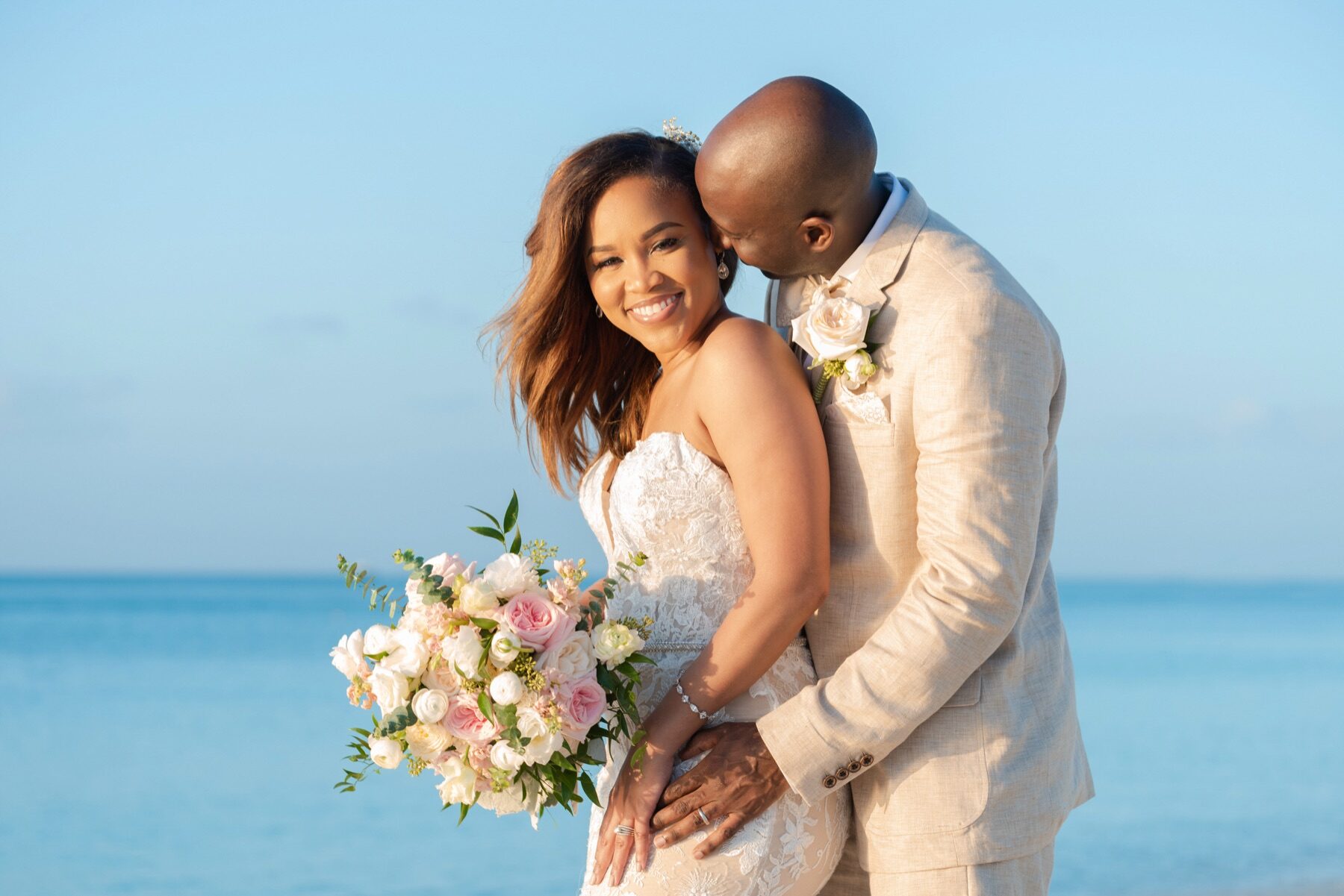 Groom in cream suit nuzzling his smiling bride's cheek against a blue sky backdrop