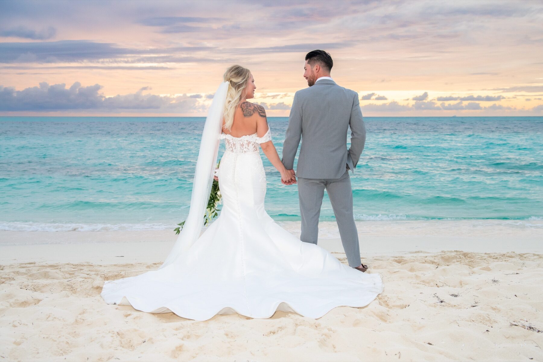 Bride and groom walking away from camera along the shore at pastel sunset