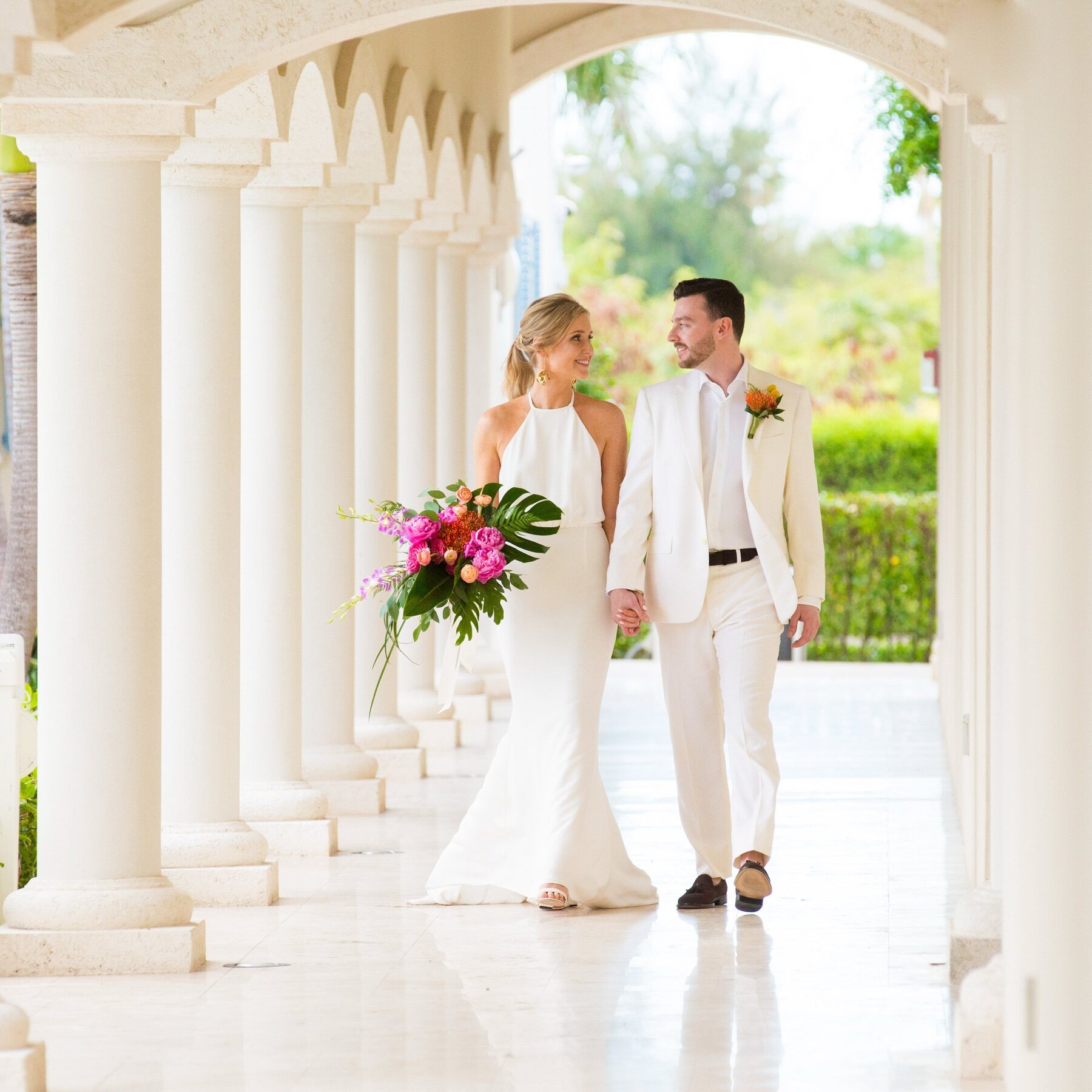Bride and groom walking arm in arm through a sunlit stone colonnade with a bright bouquet