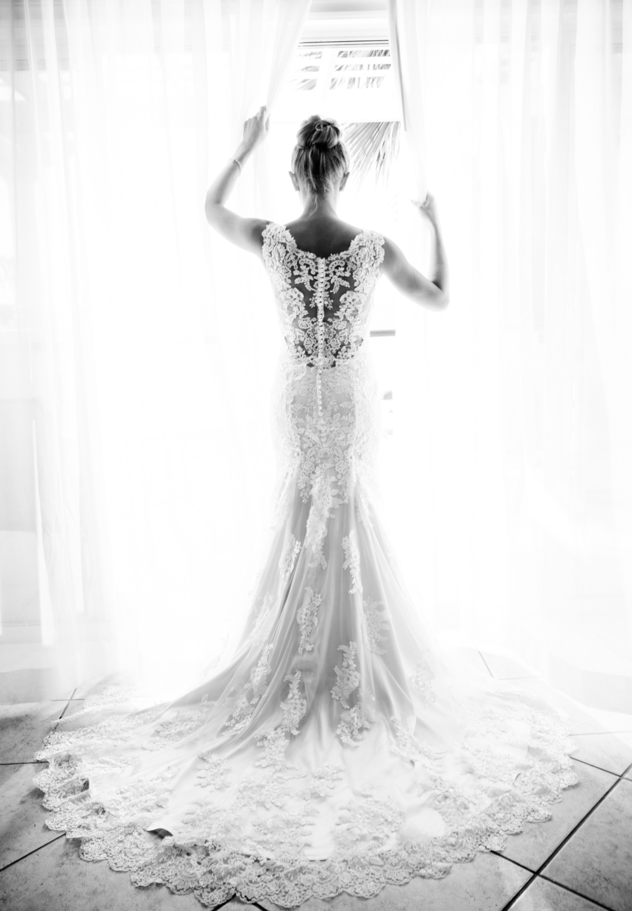 Black and white portrait of bride's lace gown from behind while facing a bright window
