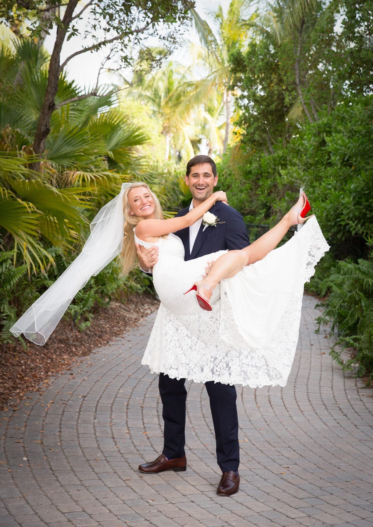 Groom carrying bride with red heels kicked up along a curved brick garden path