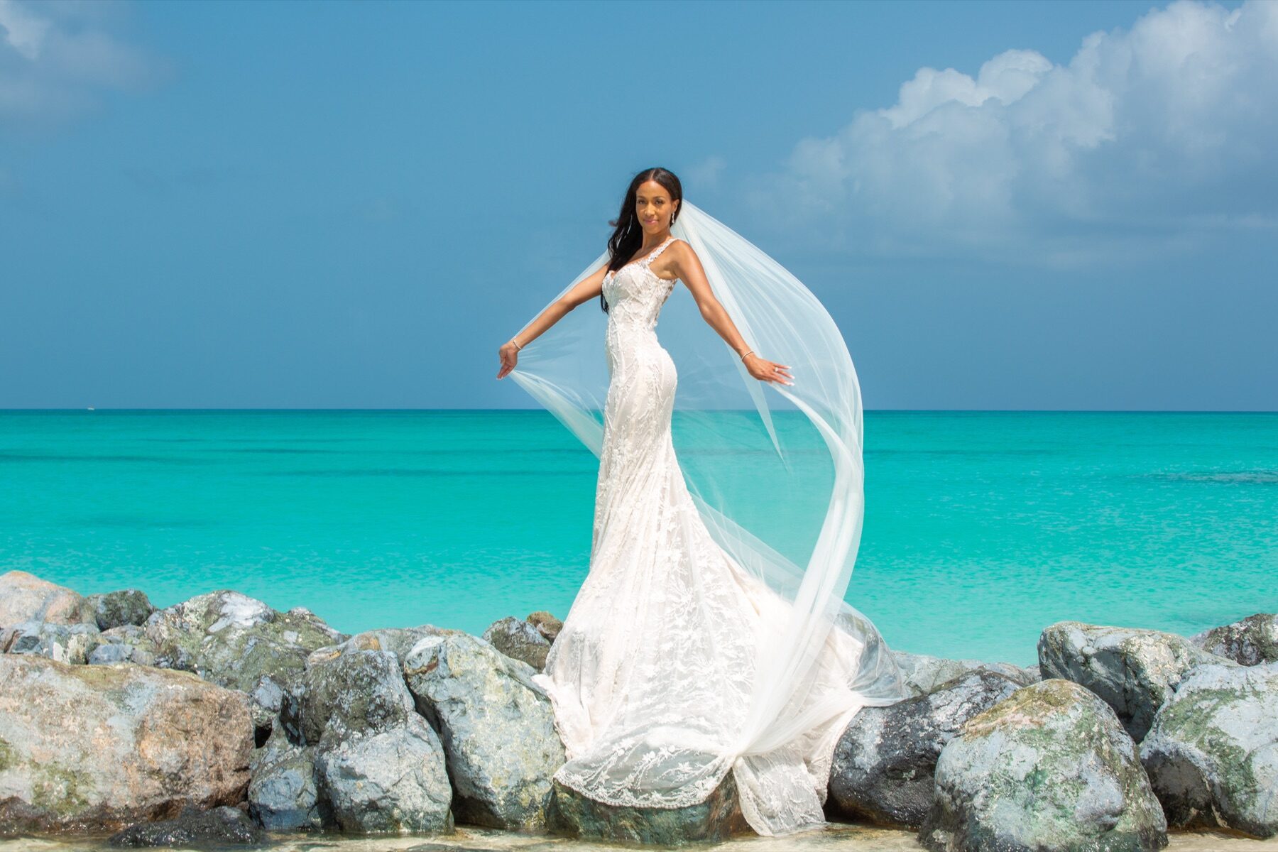 Bride twirling her veil while standing on rocks beside turquoise water