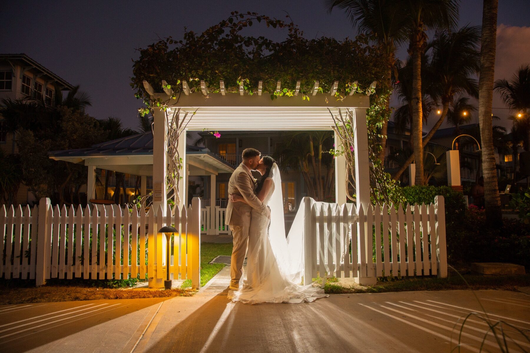 Bride and groom kissing beside a lit walkway under a flowering wooden arch at night