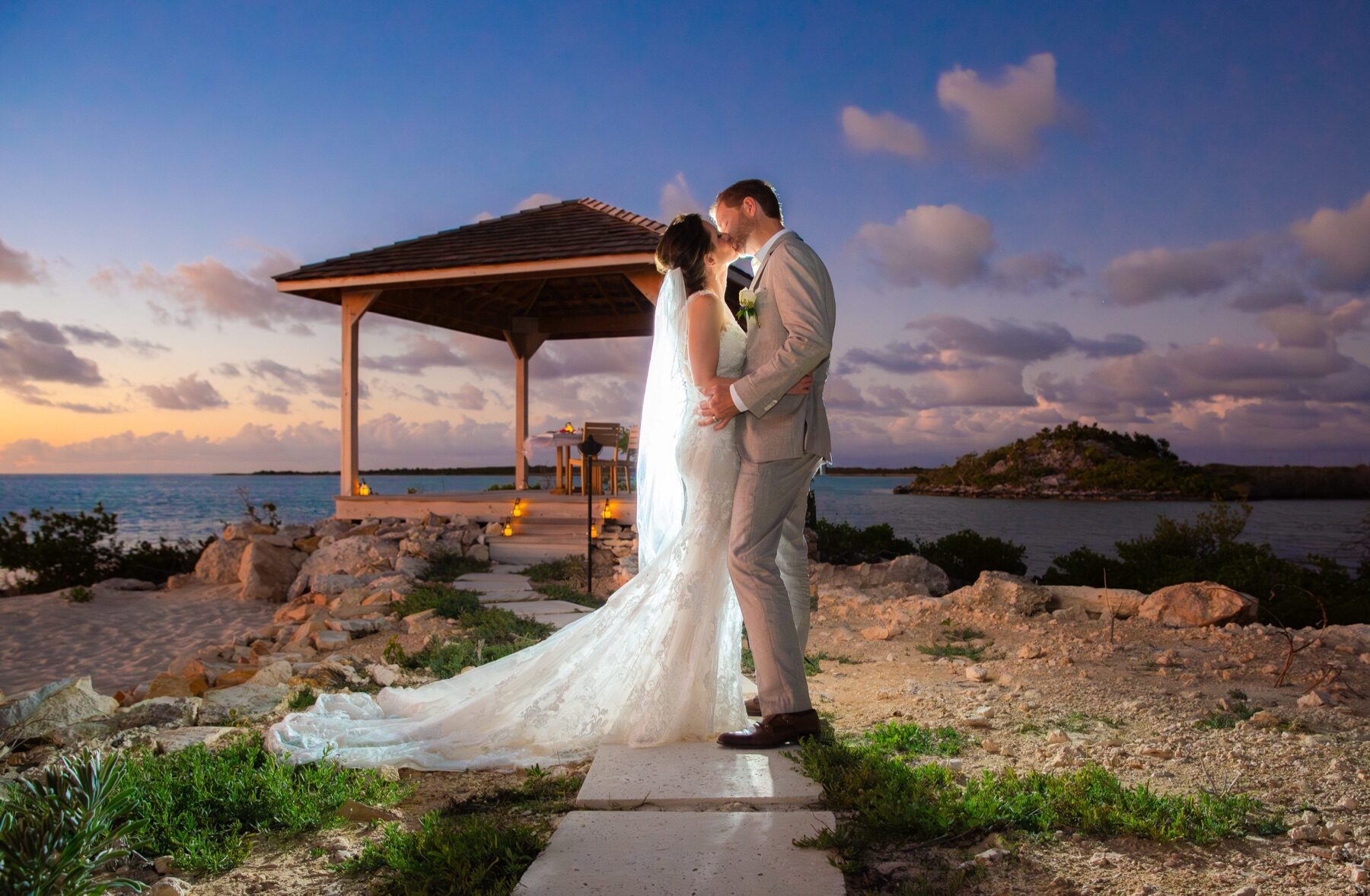 Bride and groom kissing at dusk near a waterside gazebo with an island beyond