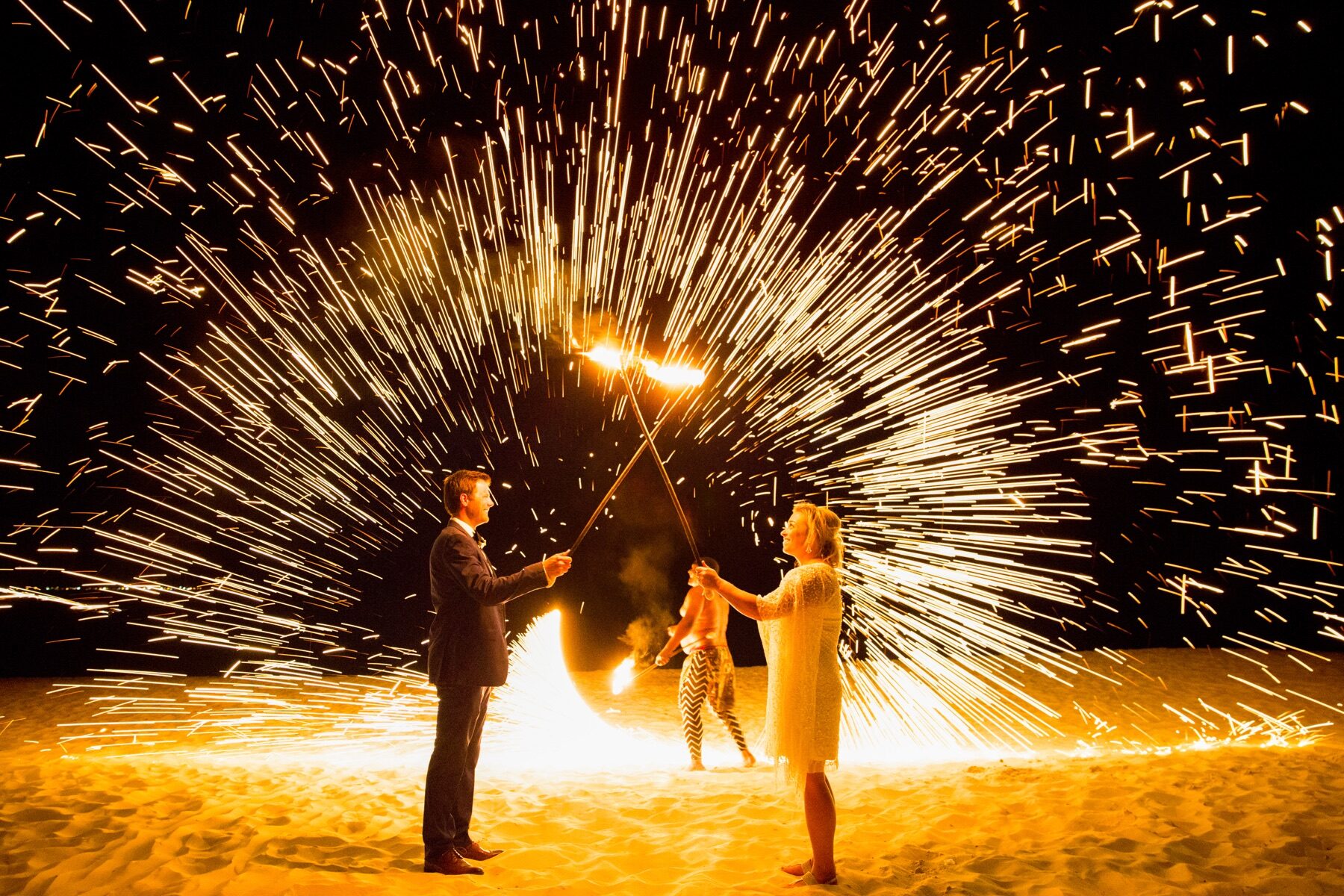 Newlyweds holding sparklers as a fire performer spins flaming poi on the sand