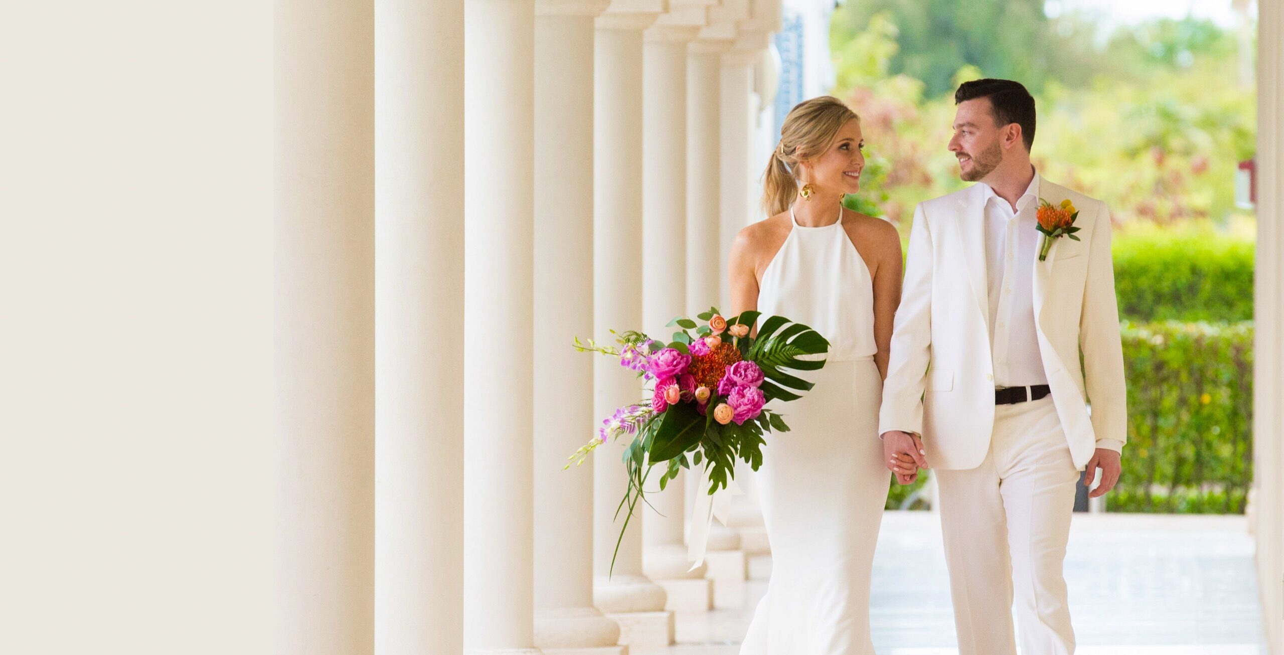 Bride and groom walking through a white colonnade with a tropical bouquet